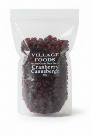 A woman in a white chef's coat holds a clear stand-up pouch of Village Foods Organic Cellared Cranberries in a kitchen setting with copper pots and tiled walls in the background.