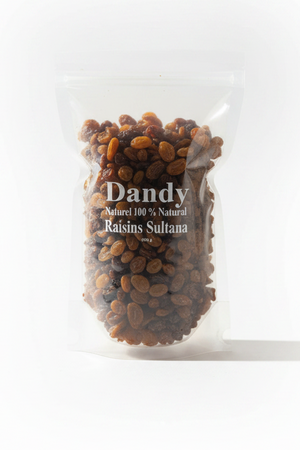 A clear stand-up pouch filled with dark raisins labeled 'Dandy Natural 100% Natural Raisins Sultana' sits on a tiled surface with a small wooden bowl of raisins in the background.
