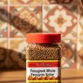 A clear plastic jar containing Fenu Greek whole seeds. The label on the jar displays the product name, weight, and indicates it as a product of Village Foods.