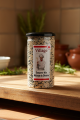 A clear plastic container labeled 'Village' with 'Sesame Mix, Mélange de Sésame' on the label, featuring an image of a chef. The container is filled with black and white sesame seeds.